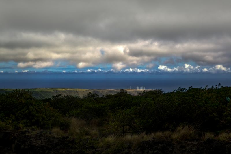 Distant wind turbines on a sunlit ridge by the ocean under heavy storm clouds