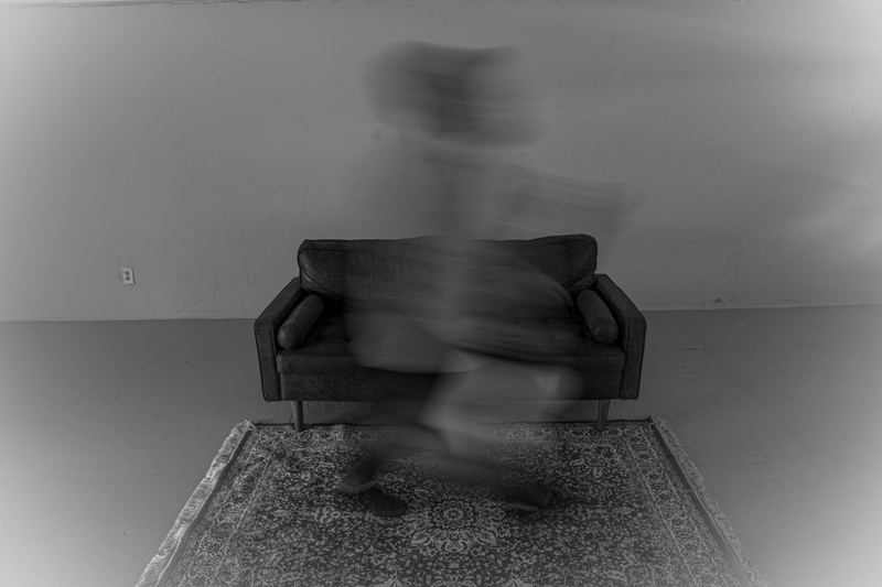 Black and white long-exposure photograph of a figure blurred in motion in front of a leather sofa on a patterned rug, an empty room behind